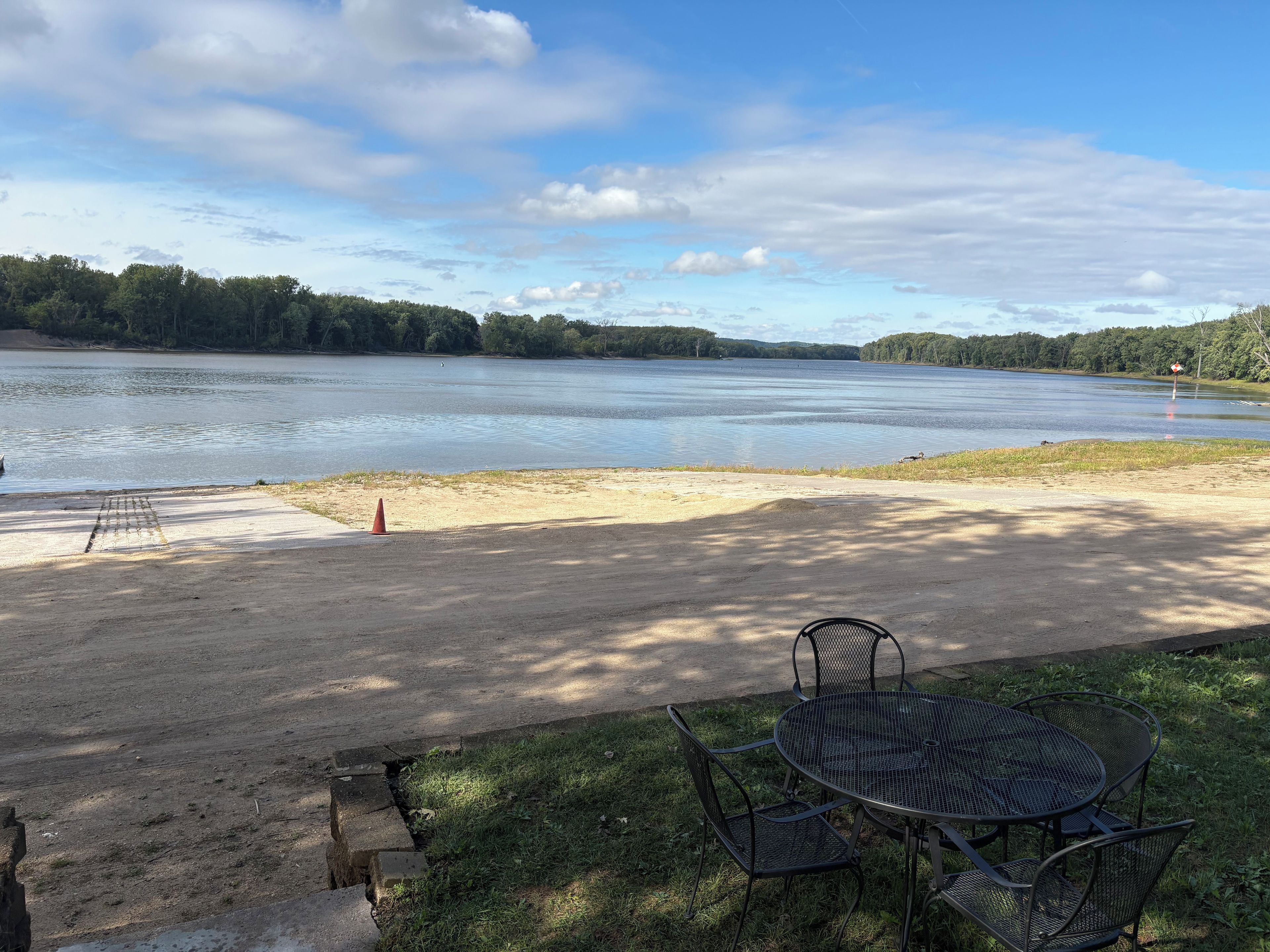 A lakeside view with a picnic table and chairs, a sandy area, and a forested background.