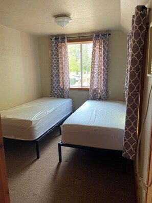Ein Zimmer mit zwei Einzelbetten, einem fensterbelaubten Fenster, einer Deckenleuchte und einem Teppichboden.