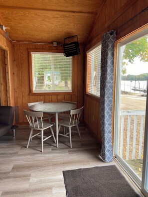 Ein Interieur einer Holzhütte mit einem Esstisch und Stühlen, einem Fenster mit Jalousien sowie Blick auf einen Steg und Wasser.