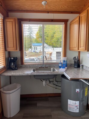 Una cocina con armarios de madera, encimera de granito, pileta de acero inoxidable, cafetera, basurero y ventana con persianas.