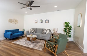 Ein Wohnzimmer mit einem blauen Sofa, einem grauen Sofa, einem grünen Sessel, einem Couchtisch und einem Deckenventilator.