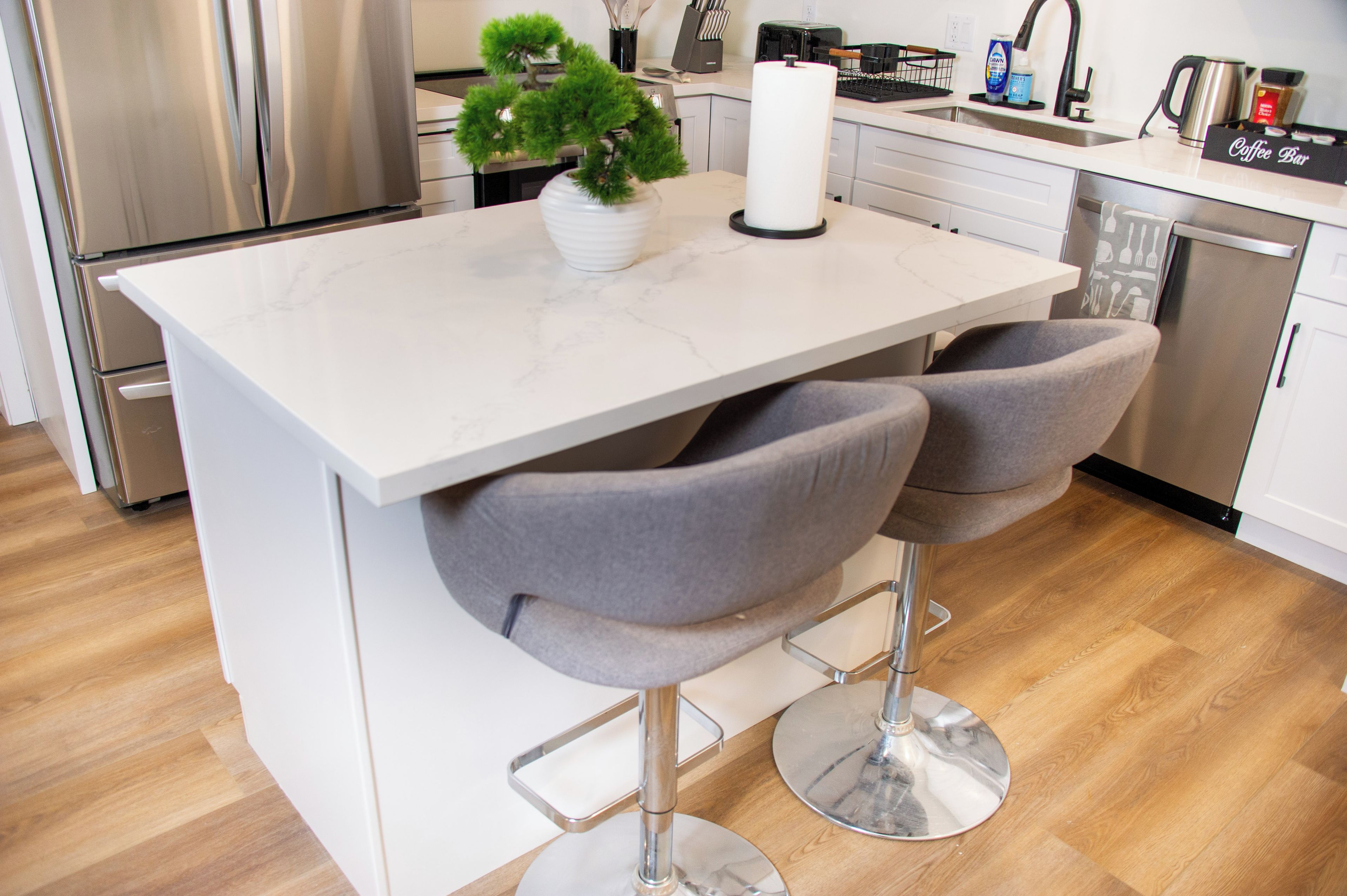 Une cuisine moderne avec un plan de travail en marbre blanc, deux tabourets de bar gris, une petite plante en pot et un coin café.