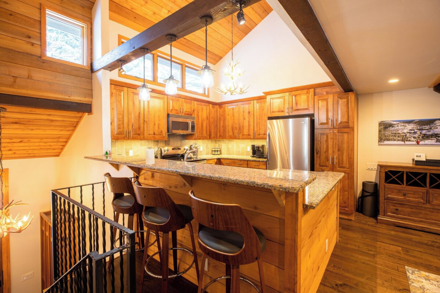A kitchen with wooden cabinets, a stainless steel refrigerator, and a bar area with stools.