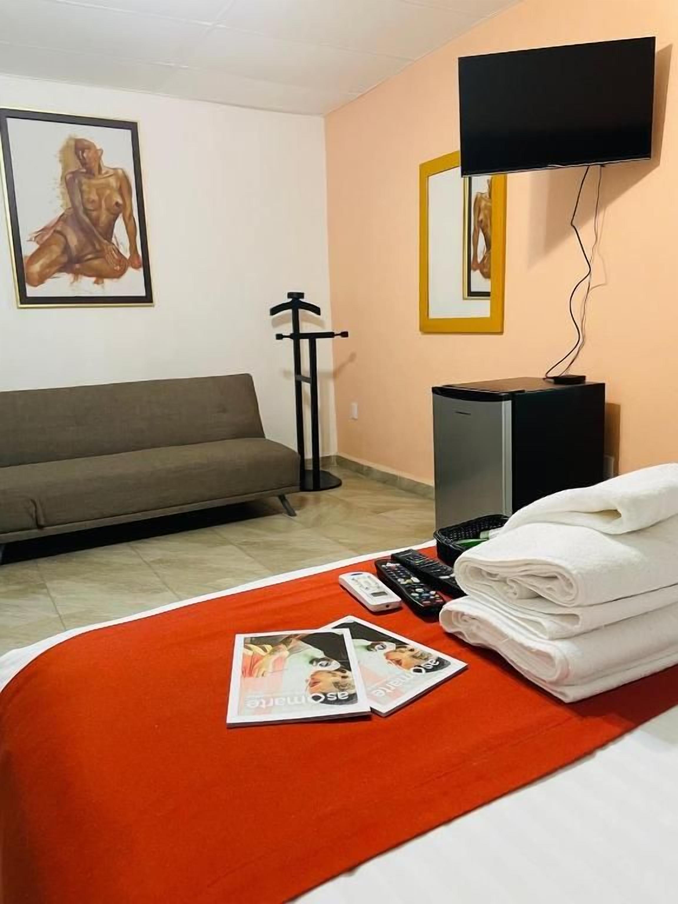 Habitación de hotel con sofá, televisor de pantalla plana montado en la pared, cama con manta roja y una pila de toallas dobladas.