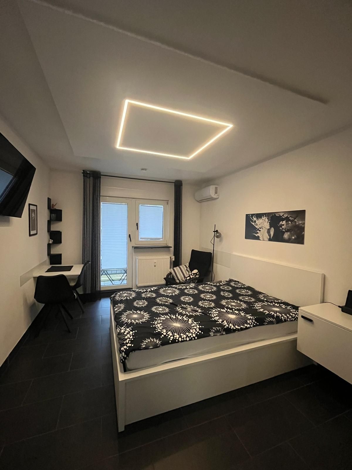 A modern bedroom with a bed, a desk, a chair, and a TV on the wall.