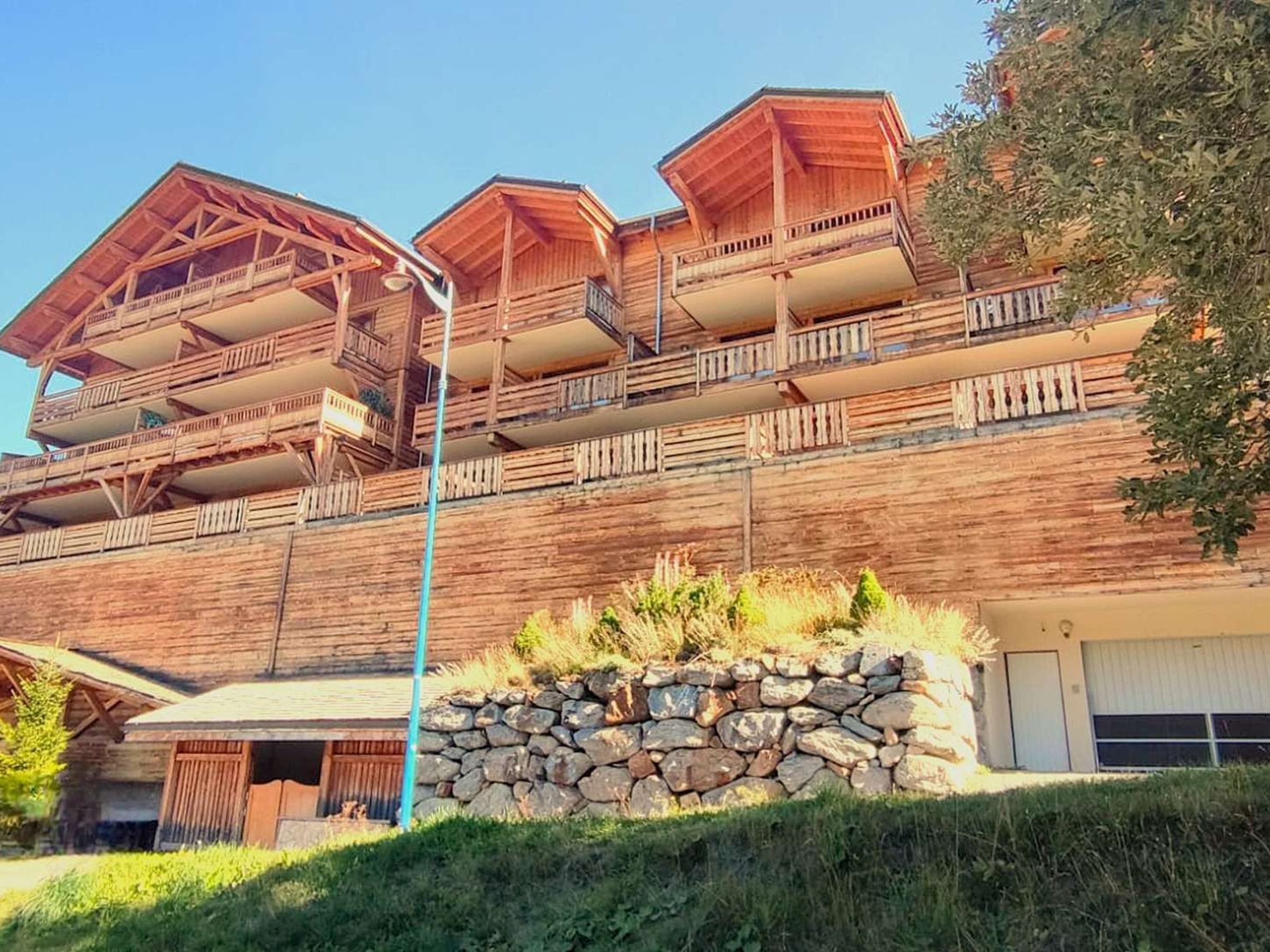 A multi-story wooden building with balconies, a stone wall in front, and a garage.