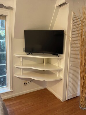 A corner room with a white entertainment unit holding a flat-screen TV, wooden flooring, and a window with blinds. - Magnolia Cottage at Saluda River Farms (Greenville)