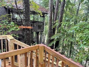 A wooden deck leading to a treehouse with a metal roof, surrounded by dense forest. - Magnolia Cottage at Saluda River Farms (Greenville)