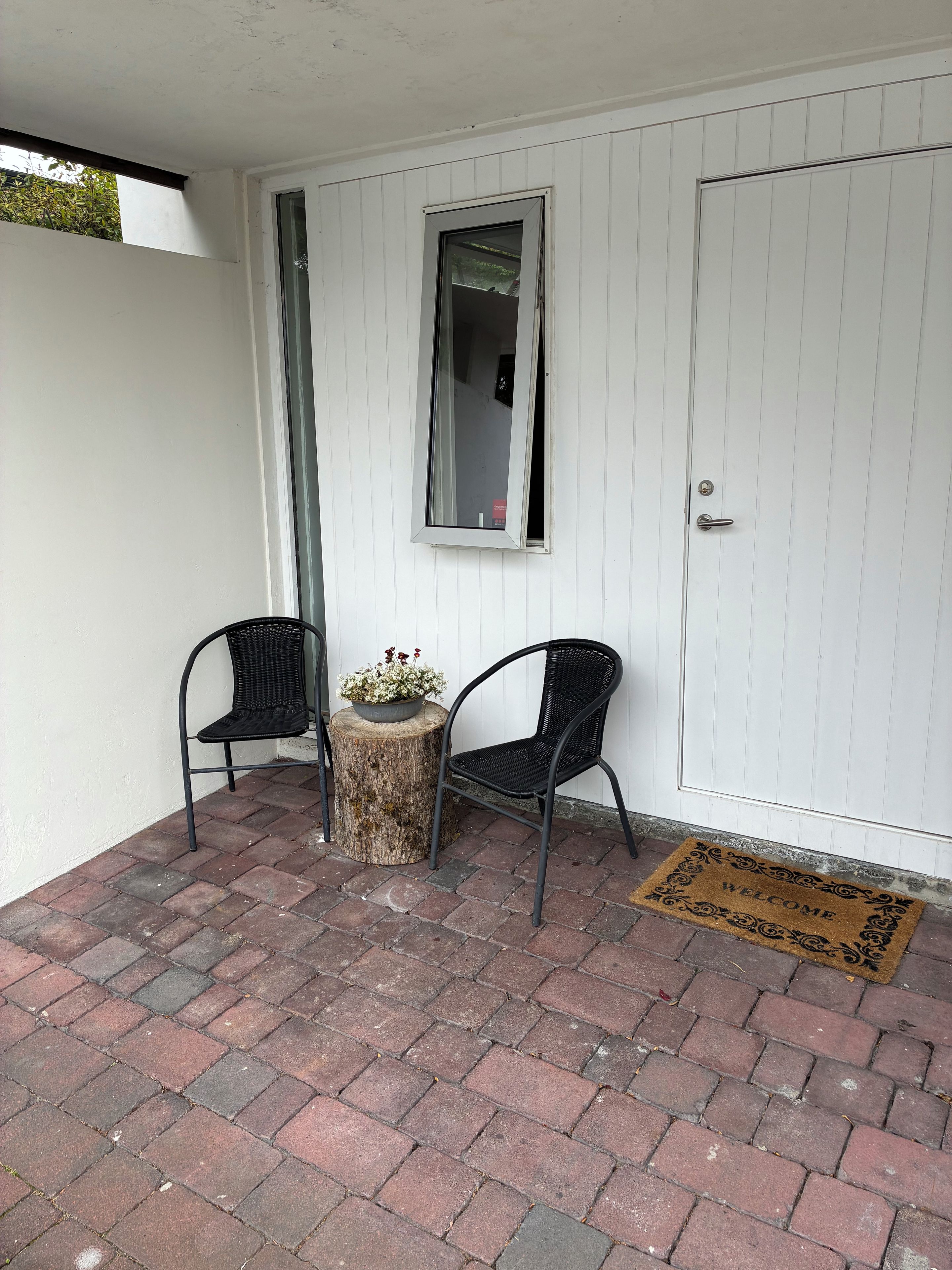A small outdoor seating area with two chairs and a table, a door with a doormat, and a window with a view of greenery.