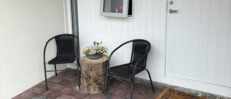 A small outdoor seating area with two chairs and a table, a door with a doormat, and a window with a view of greenery.