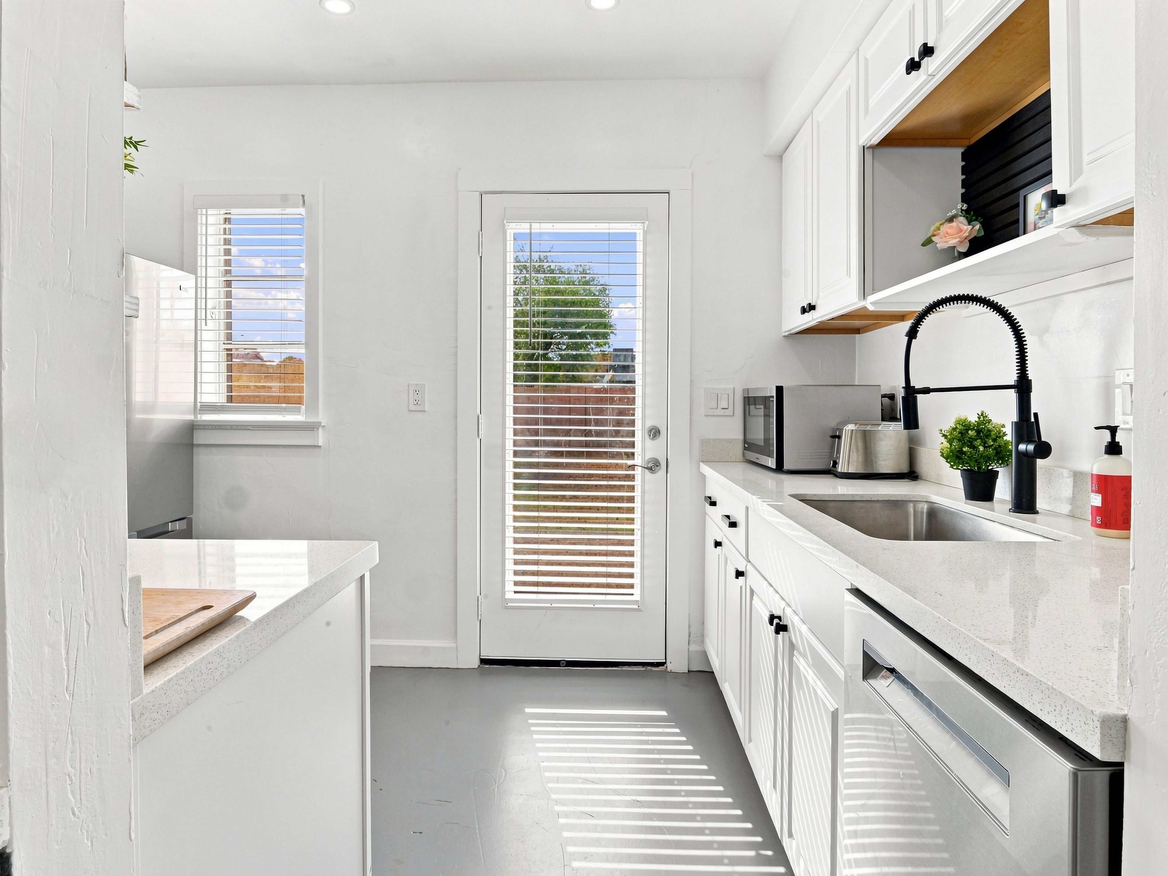 A modern kitchen with white cabinetry, a stainless steel sink, a microwave, and a toaster. There is a door with a window and blinds, and a small potted plant on the countertop.