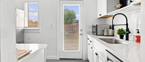 A modern kitchen with white cabinetry, a stainless steel sink, a microwave, and a toaster. There is a door with a window and blinds, and a small potted plant on the countertop.