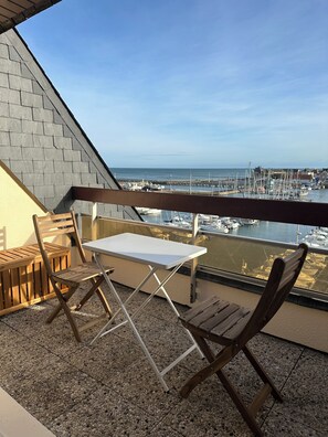 Un balcon avec une table et des chaises, donnant sur une marina où sont amarrés des bateaux et sur une jetée.