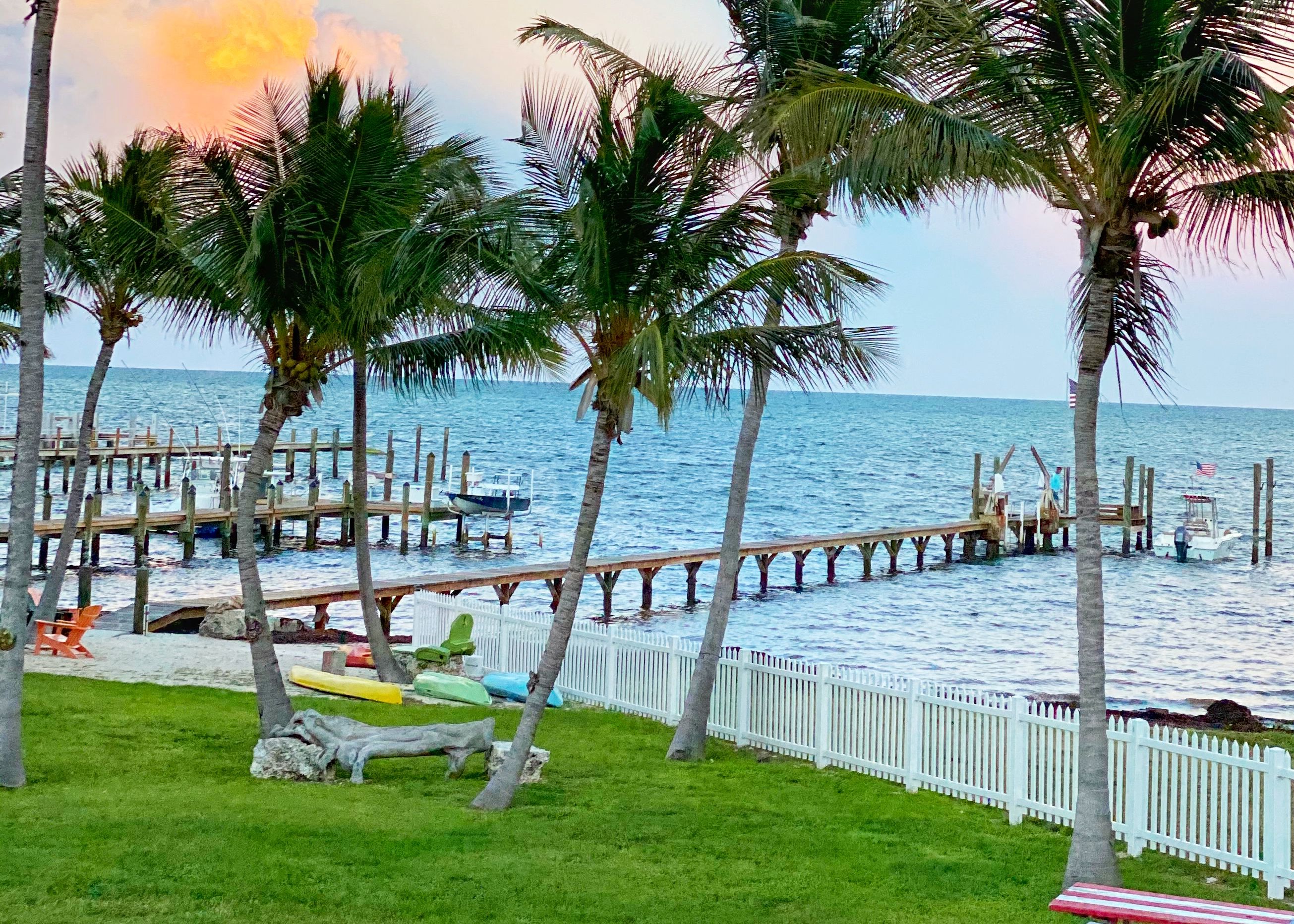 A pier extending into calm waters with palm trees and a white fence along its length.