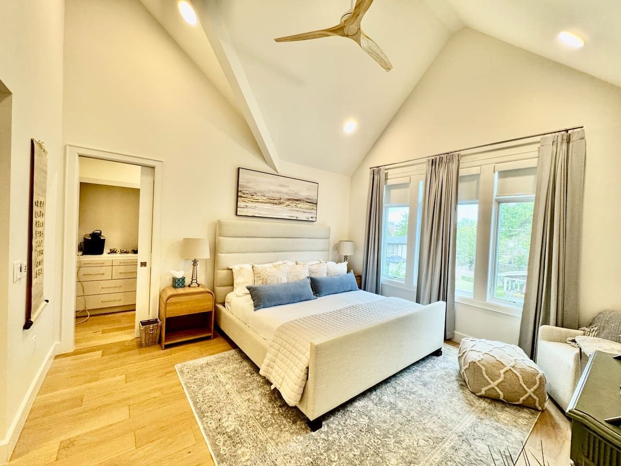 A bedroom with a bed, bedside tables, a ceiling fan, a window with curtains, and a view of greenery outside.