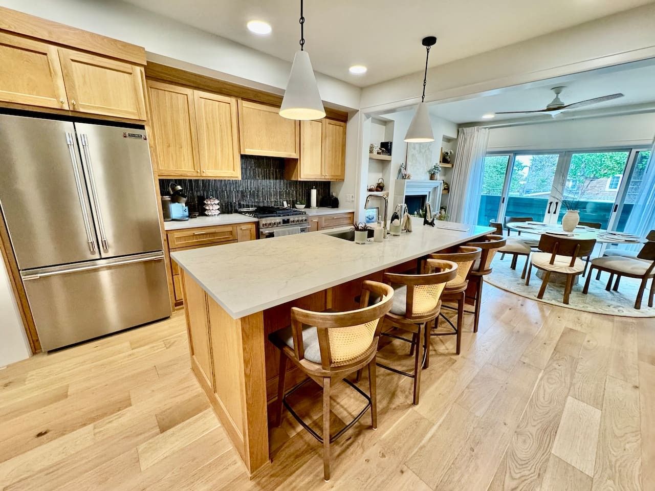 A modern kitchen with wooden cabinets, a stainless steel refrigerator, a central island with bar stools, and a dining area with chairs.