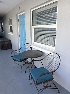 Un patio con una mesa redonda y dos sillas con cojines azules, una puerta azul y una ventana con persianas.