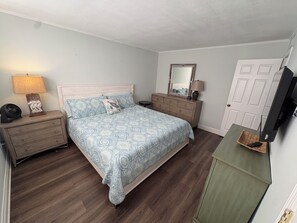 Un dormitorio con cama, mesitas de noche, cómoda, espejo, televisor y una puerta.
