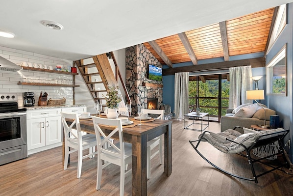A modern kitchen with white cabinets, a dining area with a wooden table, and a stone fireplace.