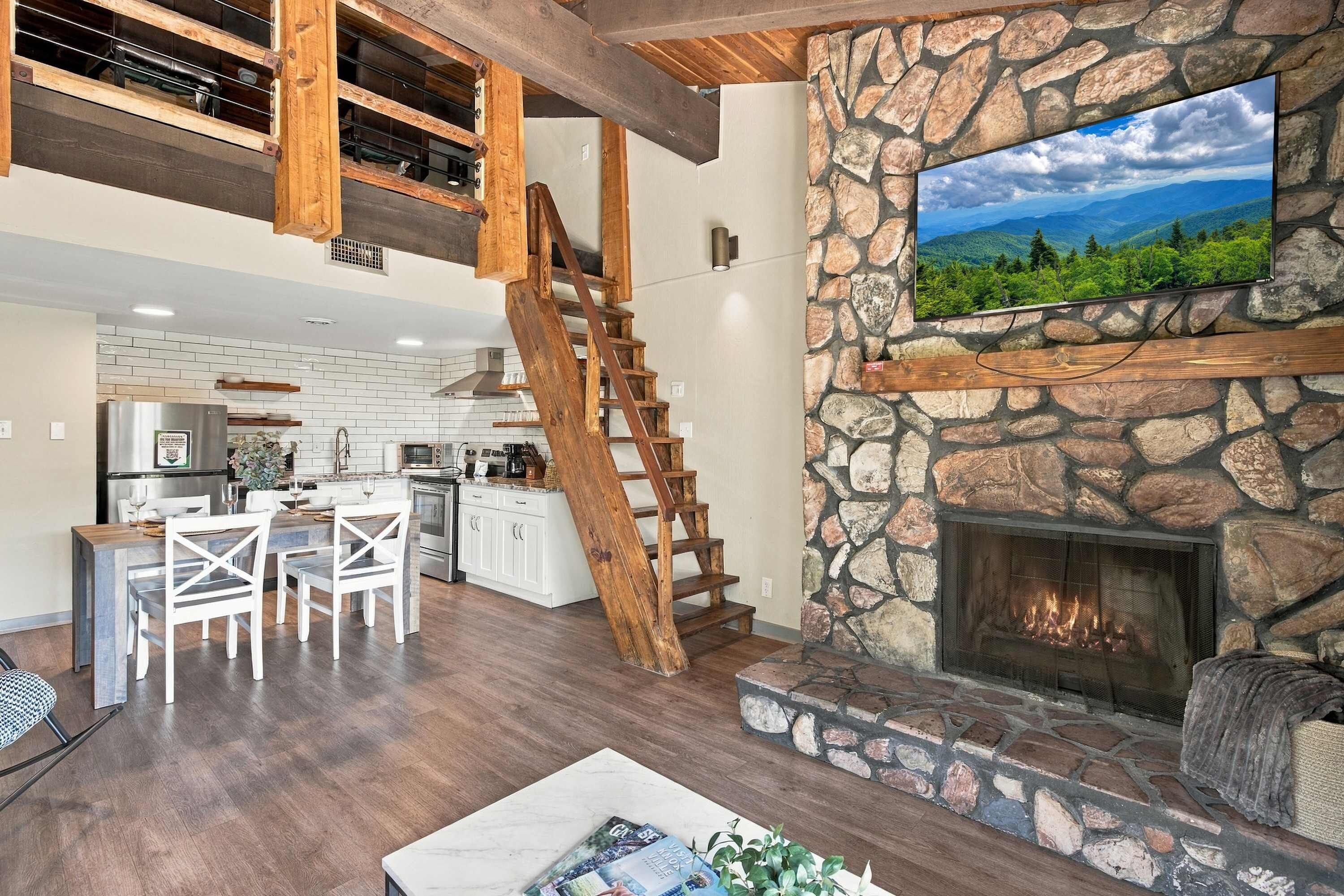 A spacious living area with a stone fireplace, a wooden staircase, and a kitchen in the background.