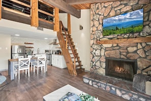 A spacious living area with a stone fireplace, a wooden staircase, and a kitchen in the background.
