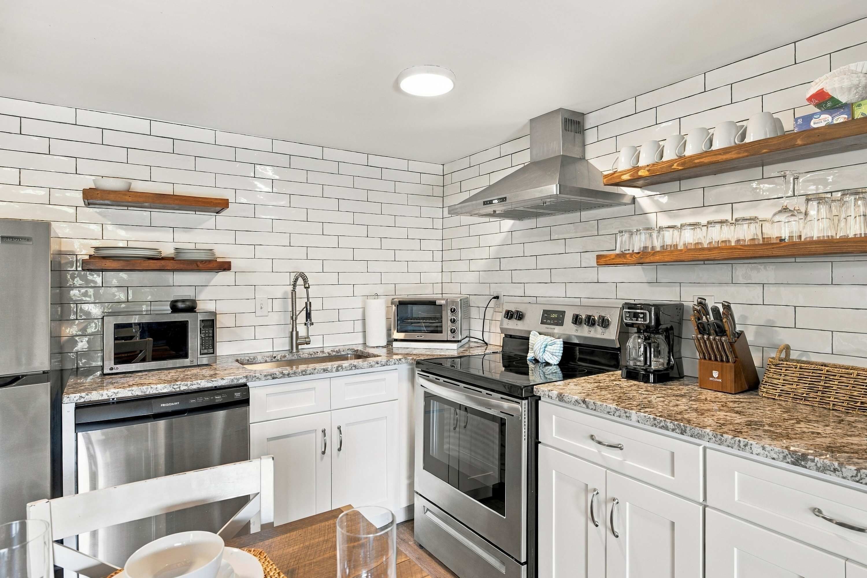A modern kitchen with white subway tiles, stainless steel appliances, wooden shelves, and a granite countertop.