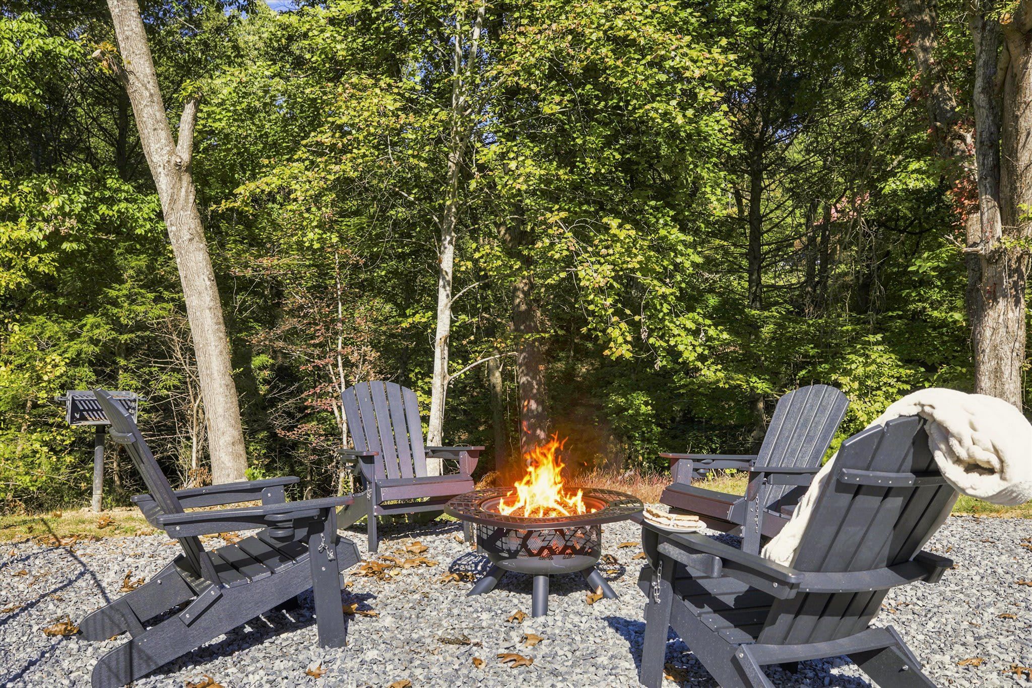 A fire pit with four Adirondack chairs arranged around it, set on a gravel surface with trees in the background.