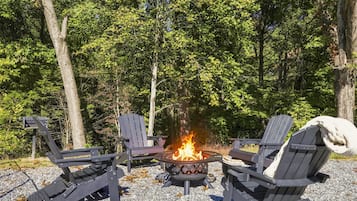 A fire pit with four Adirondack chairs arranged around it, set on a gravel surface with trees in the background.