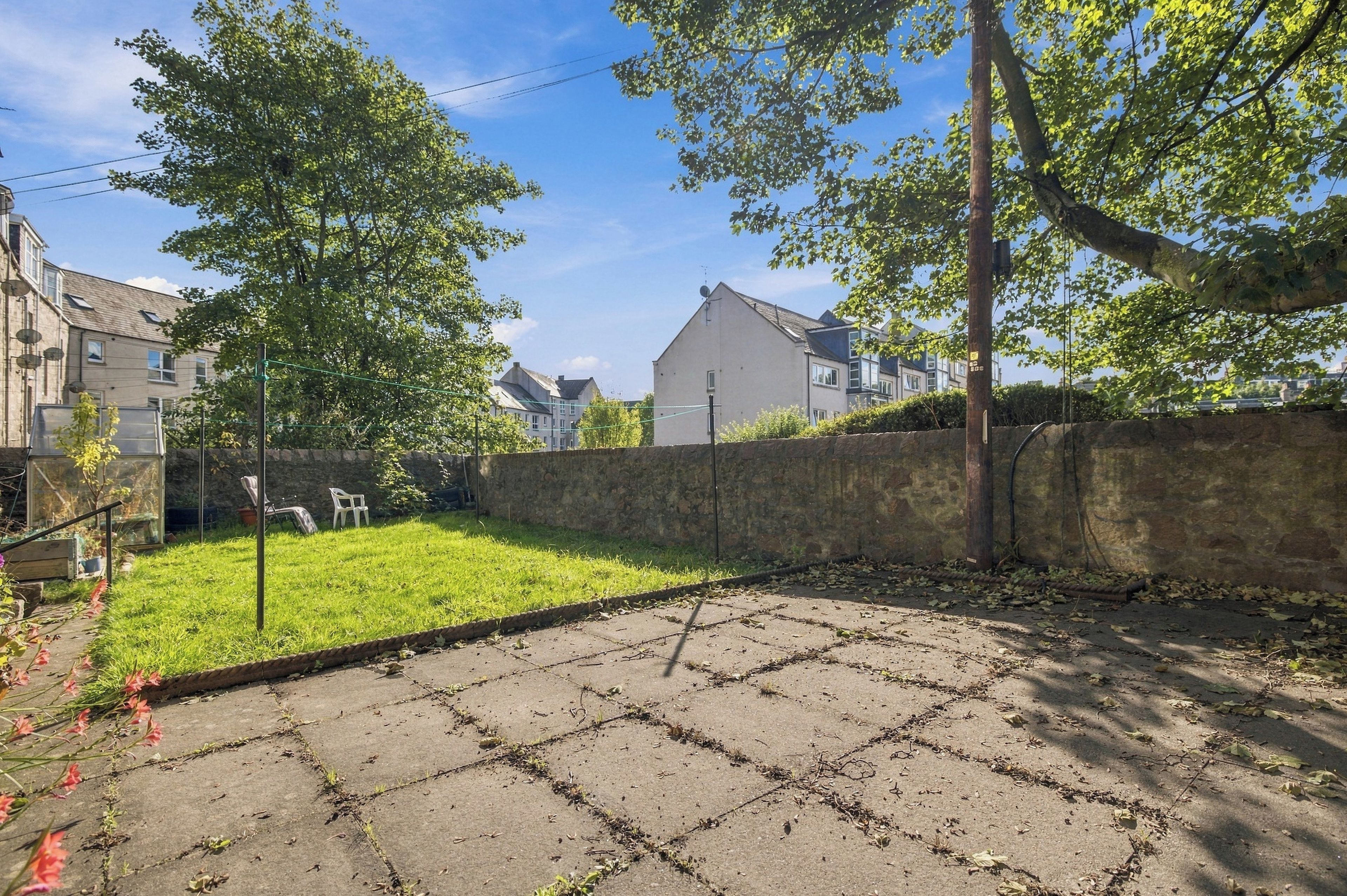 A paved courtyard with a grassy area, a stone wall, and residential buildings in the background.