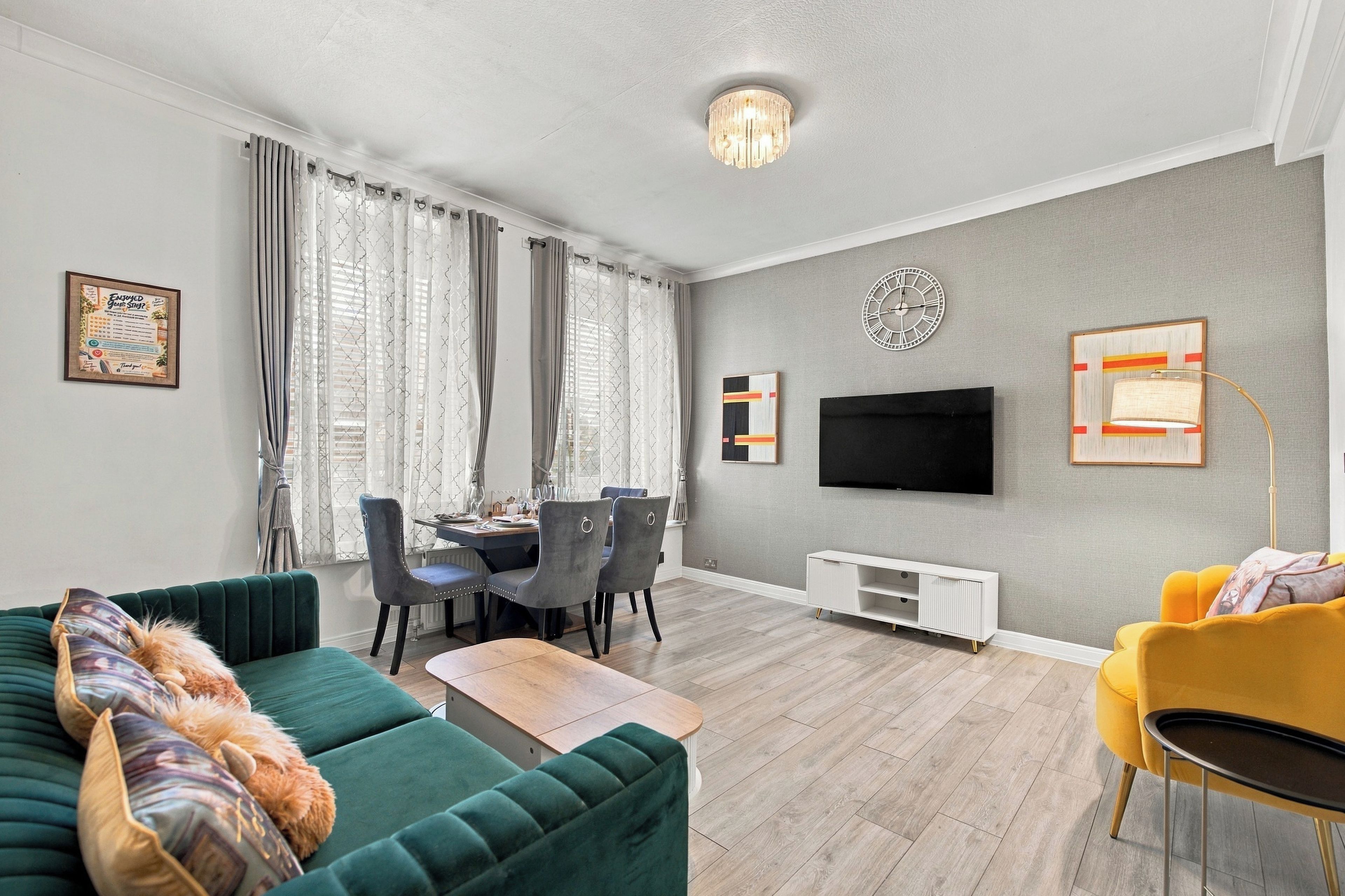 A modern living room with a green sofa, a yellow armchair, a dining area with a table and chairs, a flat-screen TV mounted on the wall, and a decorative clock.