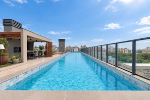 Una piscina en la azotea con vistas a la ciudad, un área de estar al aire libre techada y una fachada de edificio moderna.