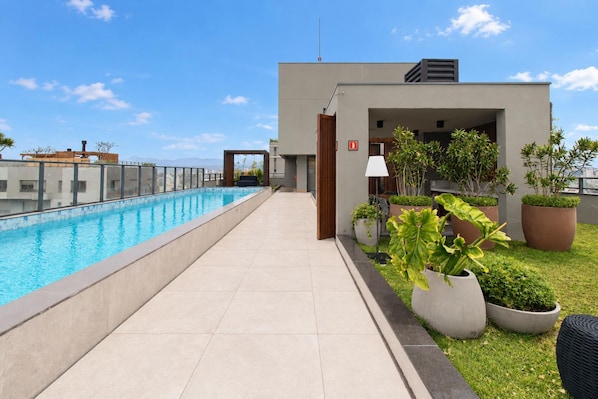 Un área de piscina en la azotea con cielo despejado, un edificio moderno de techo plano y plantas en macetas.