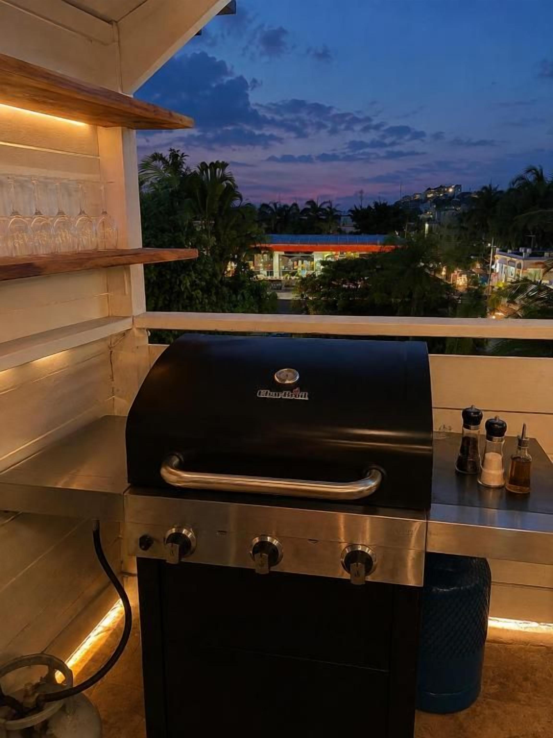 Ein Grill mit Bedienelementen, ein Metallregal mit Gläsern und ein Blick auf eine Wohngegend im Abenddämmerung.