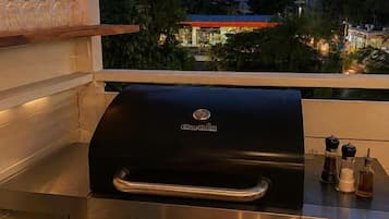 Een barbecue met bedieningselementen, een metalen plank met glazen en een uitzicht op een woonwijk in de schemering.
