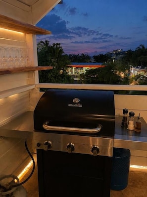 A barbecue grill with controls, a metal shelf with glassware, and a view of a residential area at dusk. - hotel Casa Roof Rooftop Beach Club  (Las Terrenas)