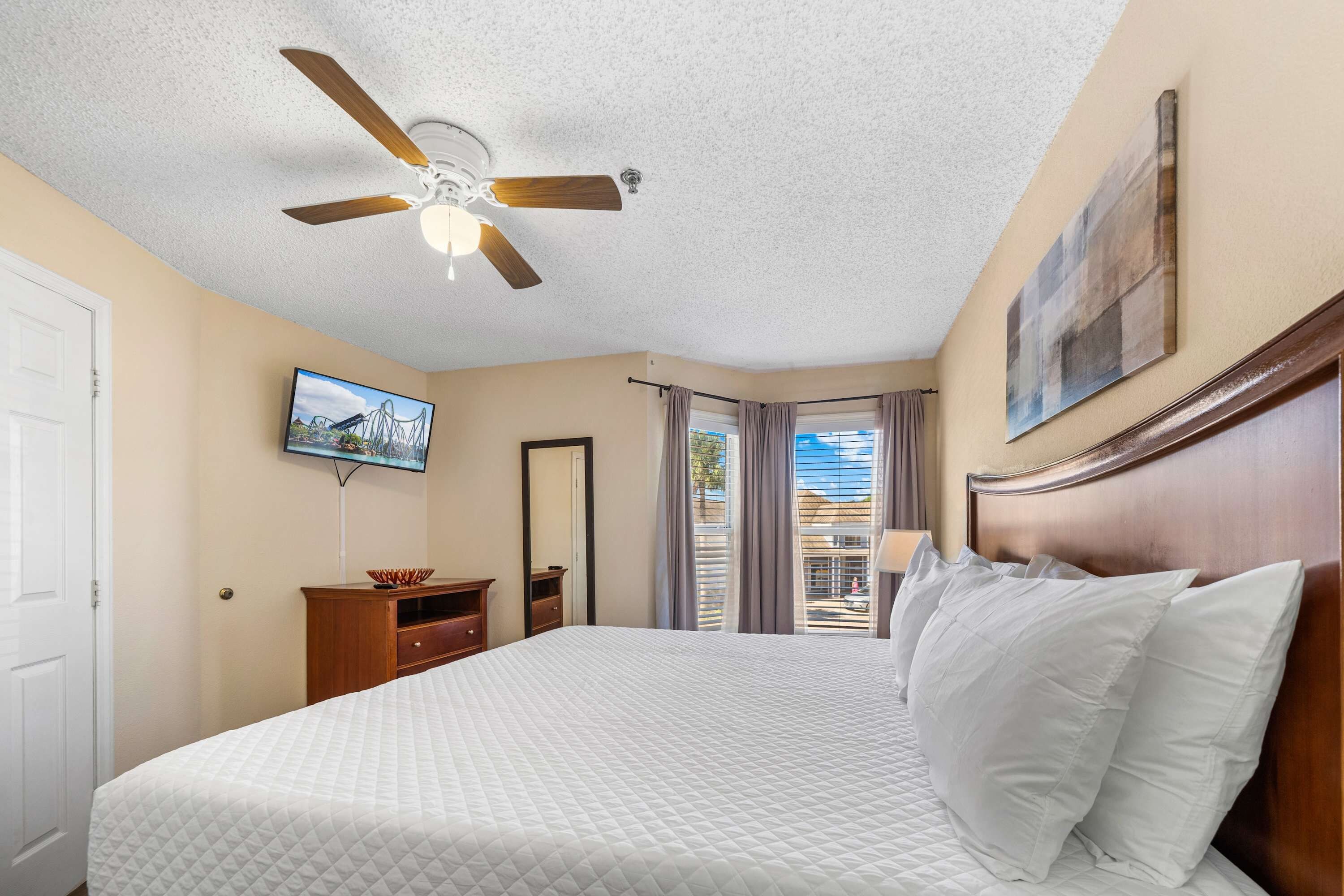 Une chambre d’hôtel équipée d’un lit, d’un ventilateur de plafond, d’une télévision, d’une commode, d’un miroir et d’une fenêtre avec des rideaux.