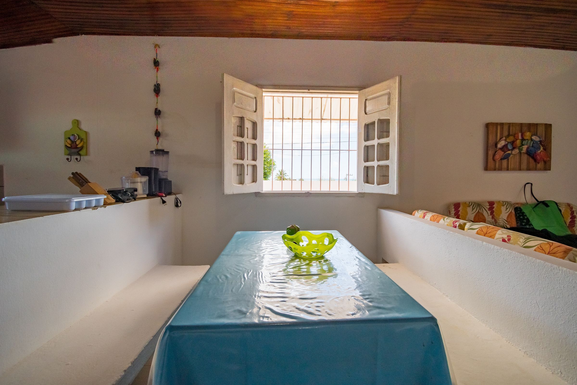 A room with a table covered by a blue tablecloth, a window with open shutters, and a wall-mounted artwork.