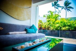 En moderne stue med en grå sofa, turkise puder, et sofabord i glas og udsigt til tropiske planter og en pool.