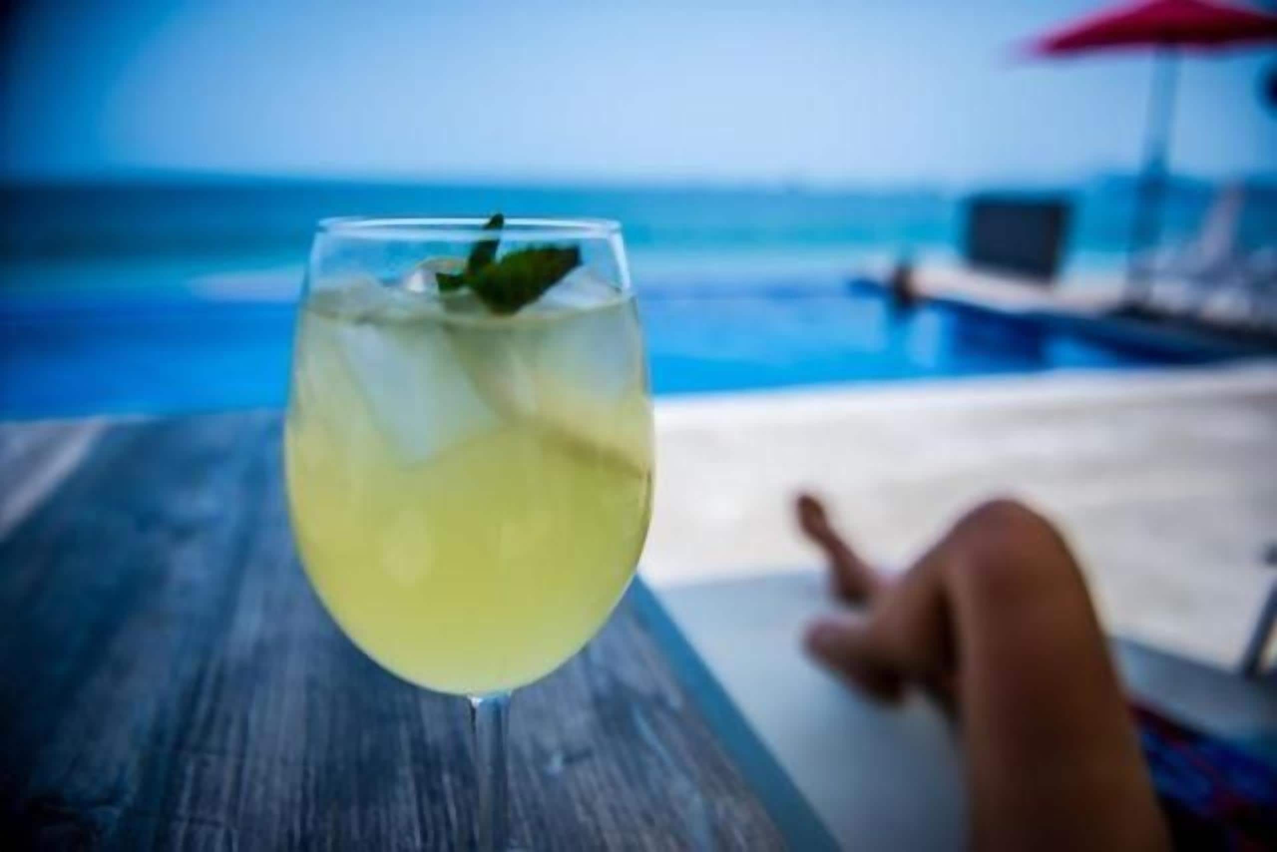 Um copo de bebida amarela com uma folha de hortelã sobre uma superfície de madeira, com um fundo desfocado de uma piscina e cadeiras de descanso.