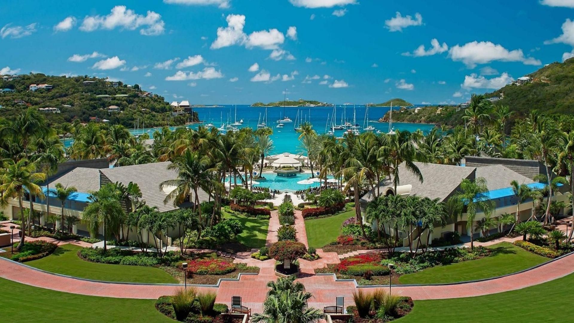 A resort with a central pool, palm trees, and a view of the ocean with boats.