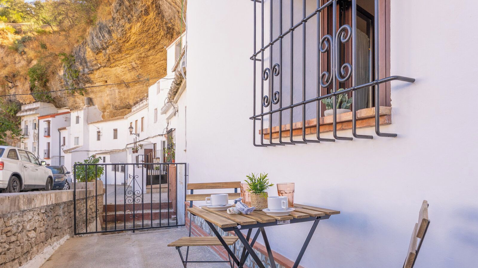 Une rue étroite avec un bâtiment blanc, une table en bois sur laquelle se trouvent des tasses et une plante, et une voiture garée à l’extérieur.