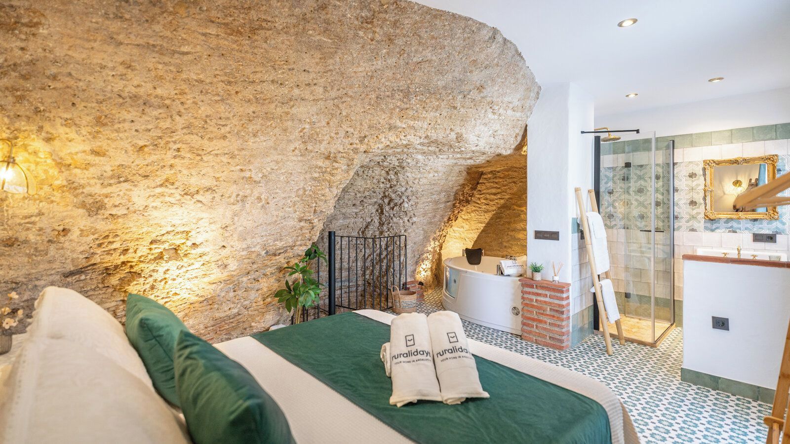 Une chambre avec un grand mur en pierre, un lit avec une literie verte, une petite table avec une lampe et une salle de bain avec une douche en verre.