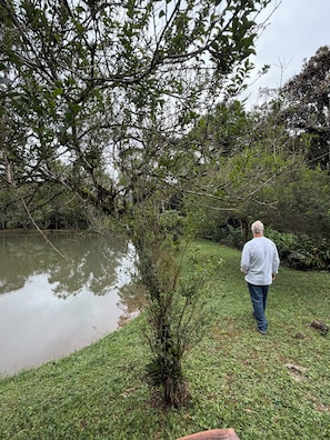 A man walking along a grassy path beside a calm body of water, surrounded by dense foliage. - Estância 3 por 1 (Guaratuba)