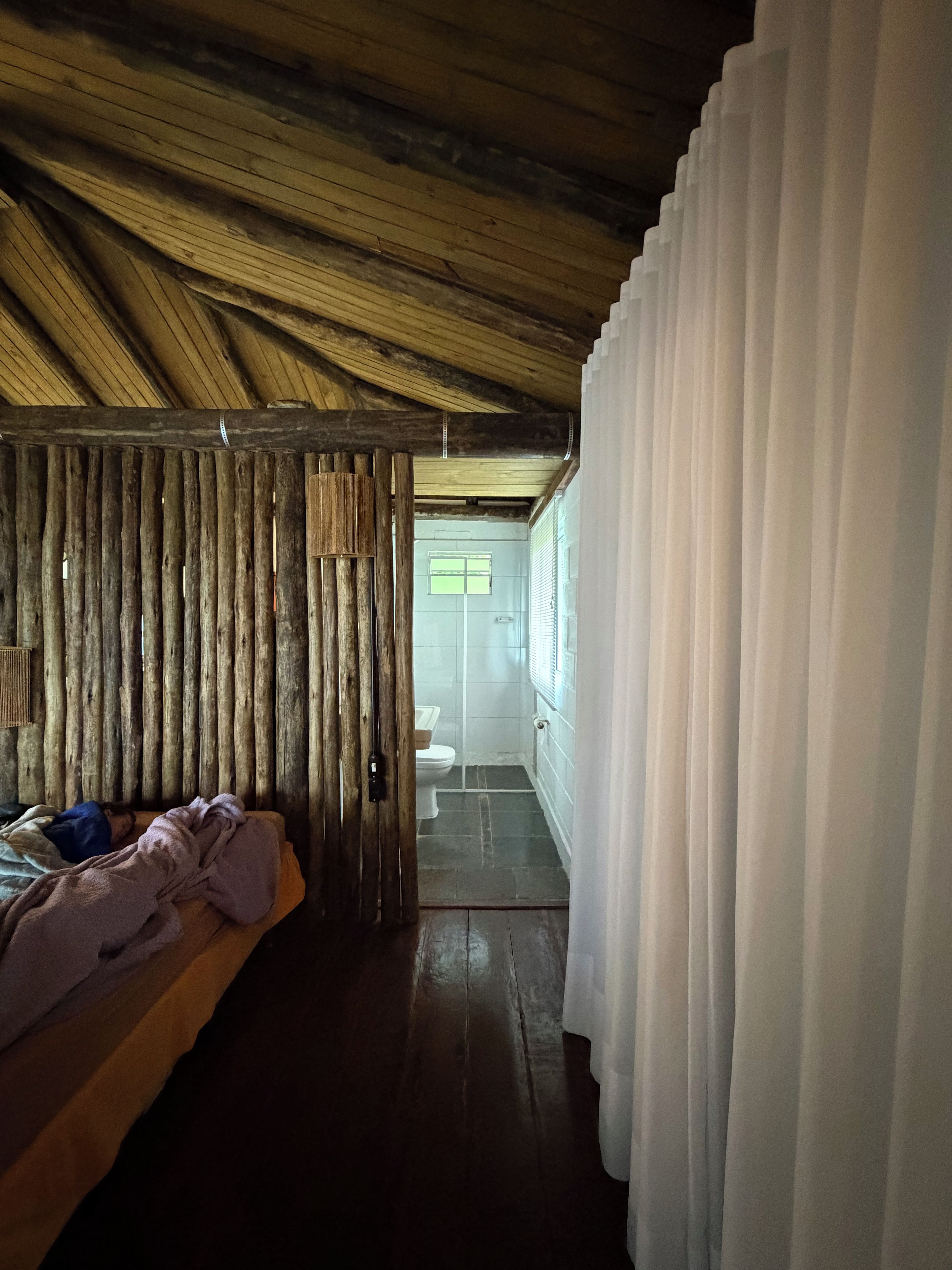 Una habitación con techo de madera, una cama con manta, un panel de madera y vistas a un baño a través de una puerta.