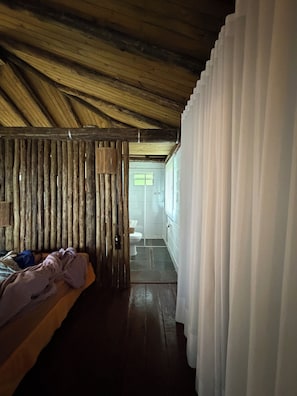 Una habitación con techo de madera, una cama con manta, un panel de madera y vistas a un baño a través de una puerta.