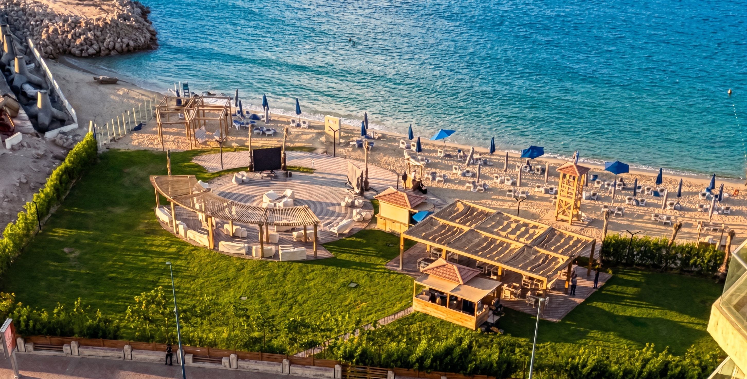 Vue aérienne d’un complexe hôtelier en bord de mer, avec un espace de détente circulaire, des chaises longues et un petit bâtiment.
