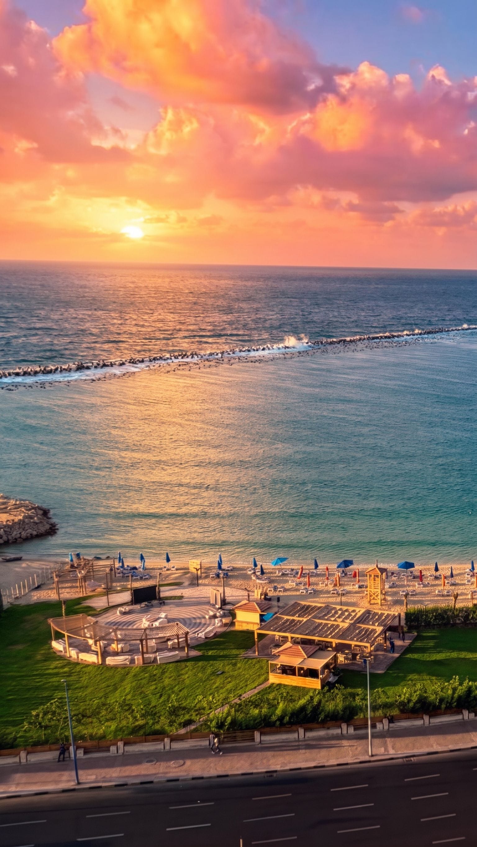 A coastal resort with a beach, sun loungers, umbrellas, and a view of the ocean at sunset.