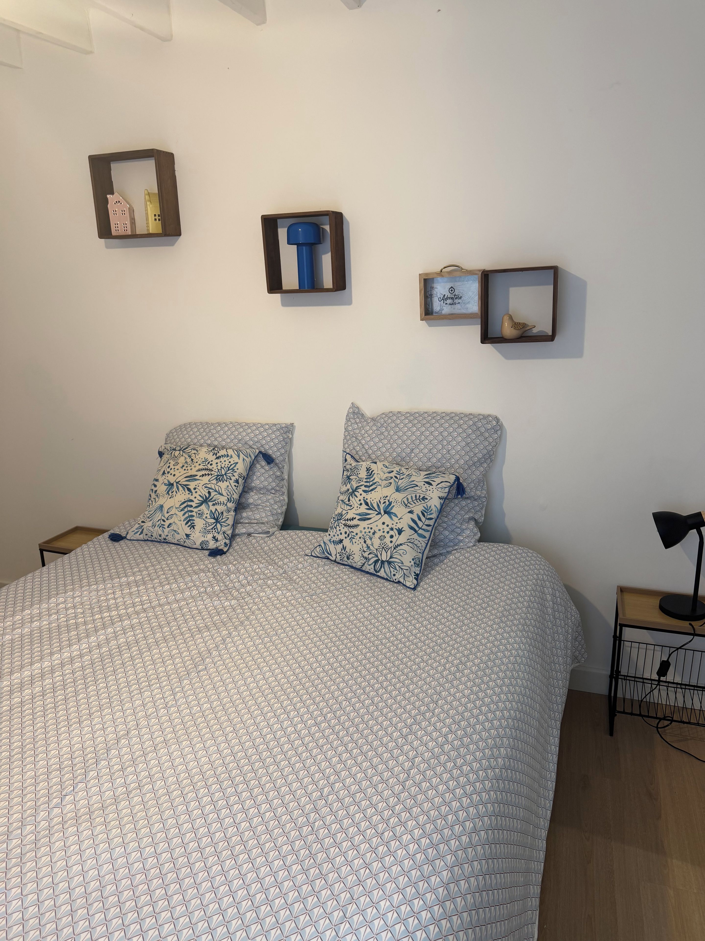 A bedroom with a bed, two pillows, a bedside table with a lamp, and wall-mounted shelves with decorative items.