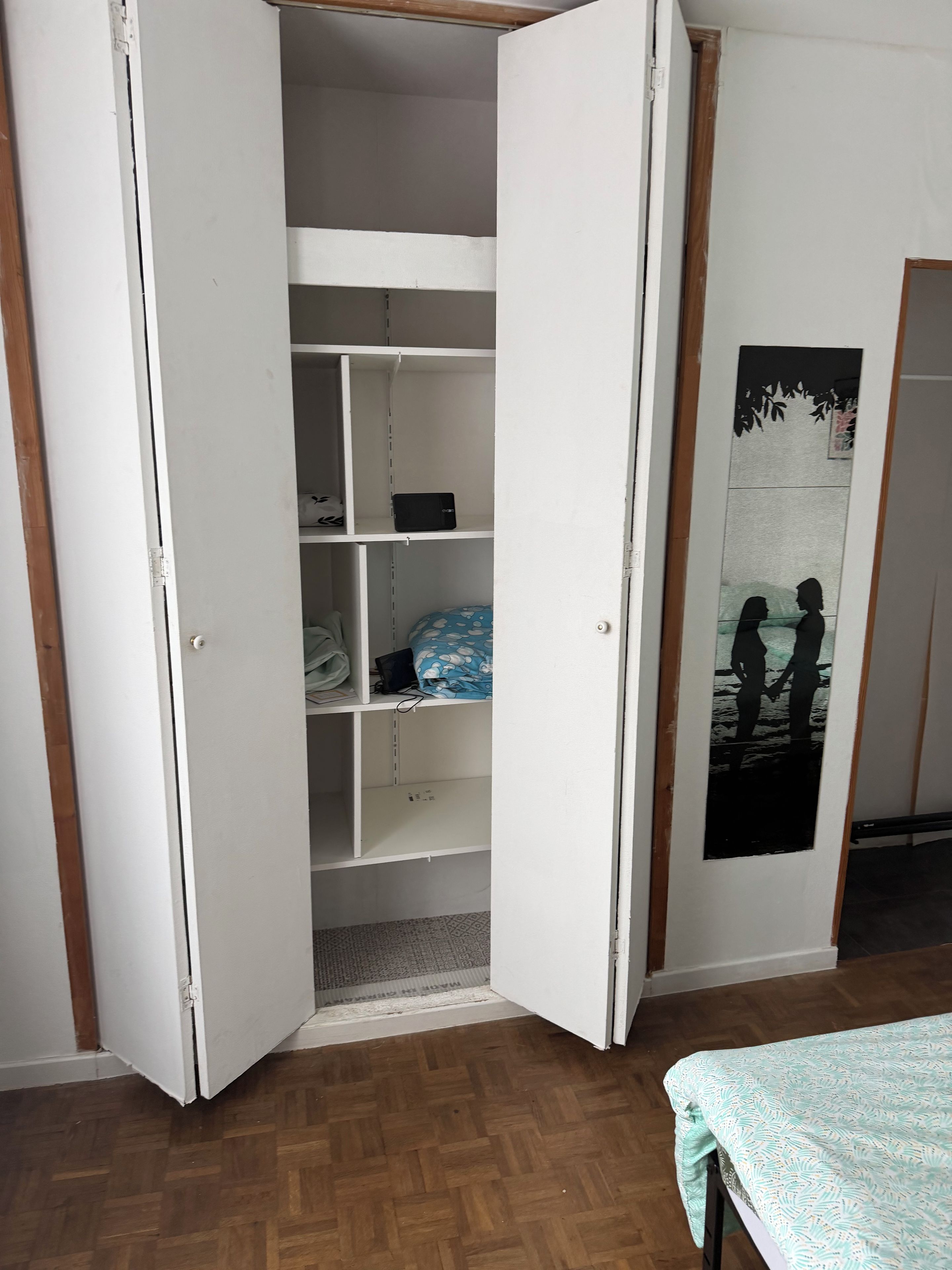 A room with a wooden floor, a bed with a patterned bedspread, and a white wardrobe with open doors revealing shelves and hanging items.
