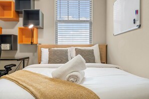 Un letto rifatto con un copriletto bianco e due cuscini decorati, un comodino con una lampada, e una lavagna applicata alla parete con strumenti di scrittura.